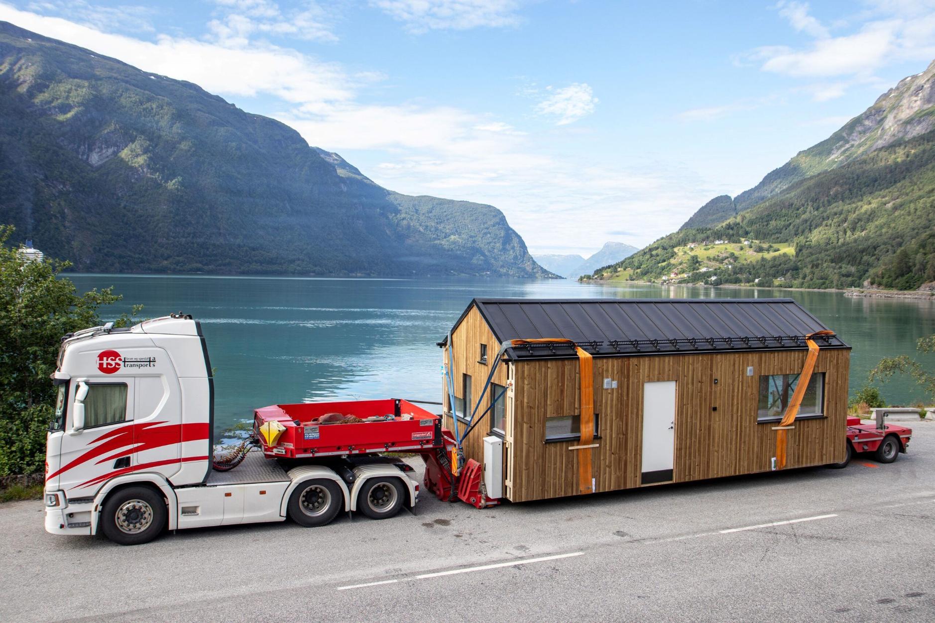 Lastebil frakter et lite hus på tilhenger langs en innsjø med fjell i bakgrunnen.