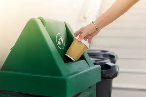 Hånd som kaster et papirkopp-lokk i en grønn søppelkasse